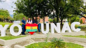 Mirian estudiante de la Universidad Adventista de Bolivia haciendo su intercambio en la UNAC junto con dos amigas sosteniendo la bandera de Bolivia en el monumento "Yo amo UNAC"